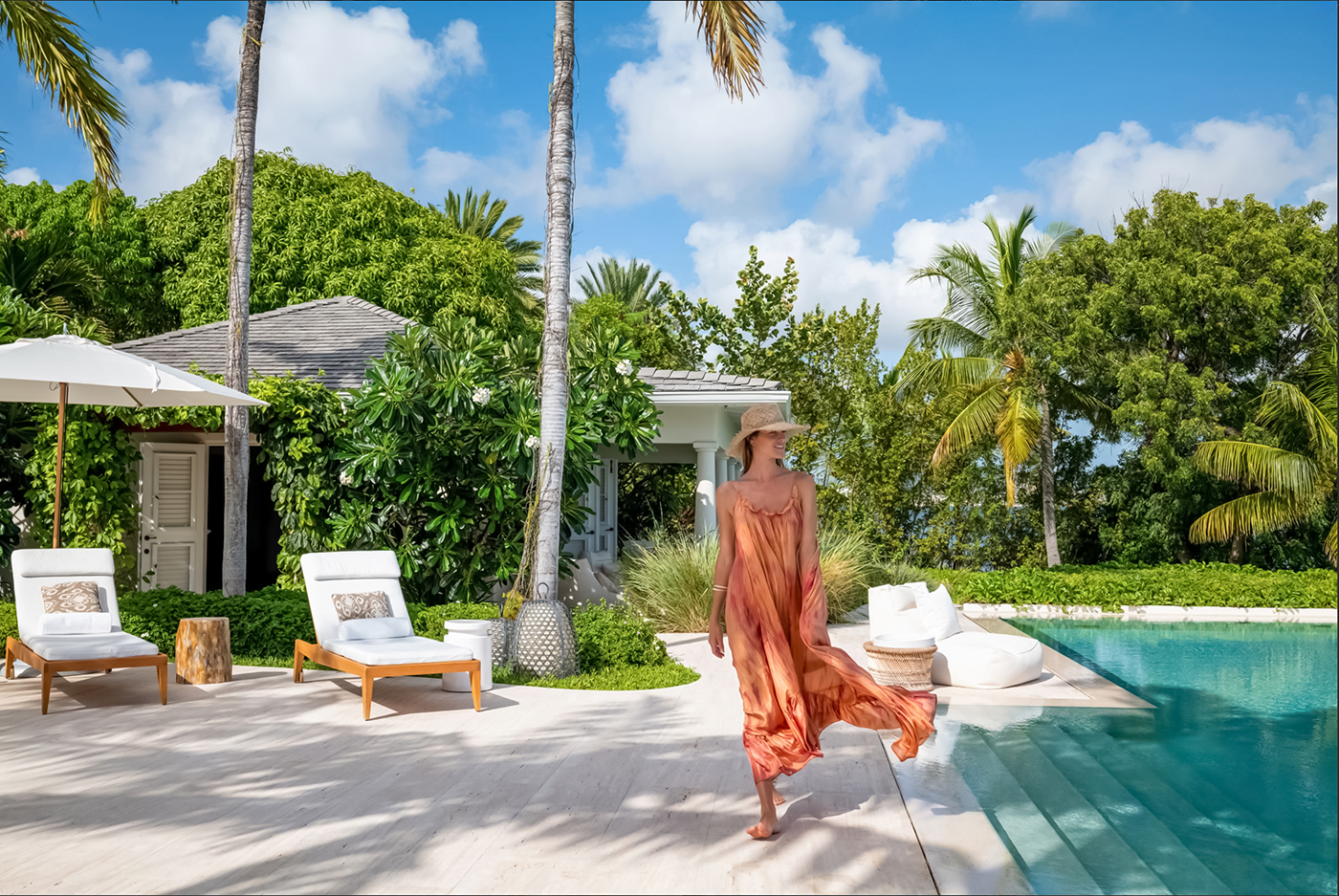 Woman in orange dress and sunhat walking barefoot beside a pool at a tropical villa with lounge chairs and lush greenery.