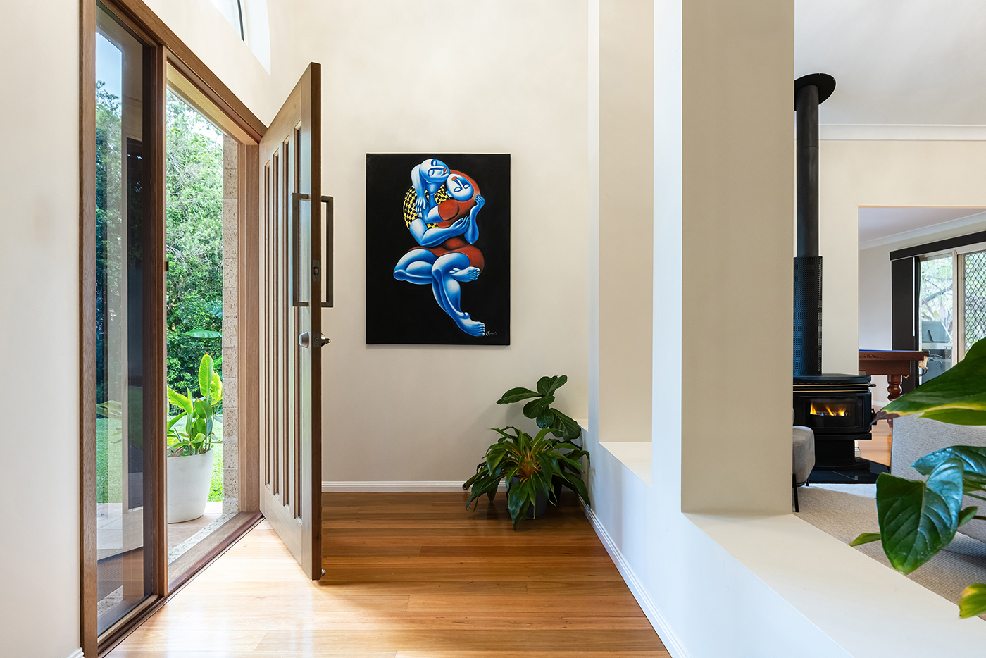 Entryway with wooden door open, wooden floor, a colorful painting of two embracing figures on the wall, and green plants near a white half wall with a black wood stove visible in the adjoining room.