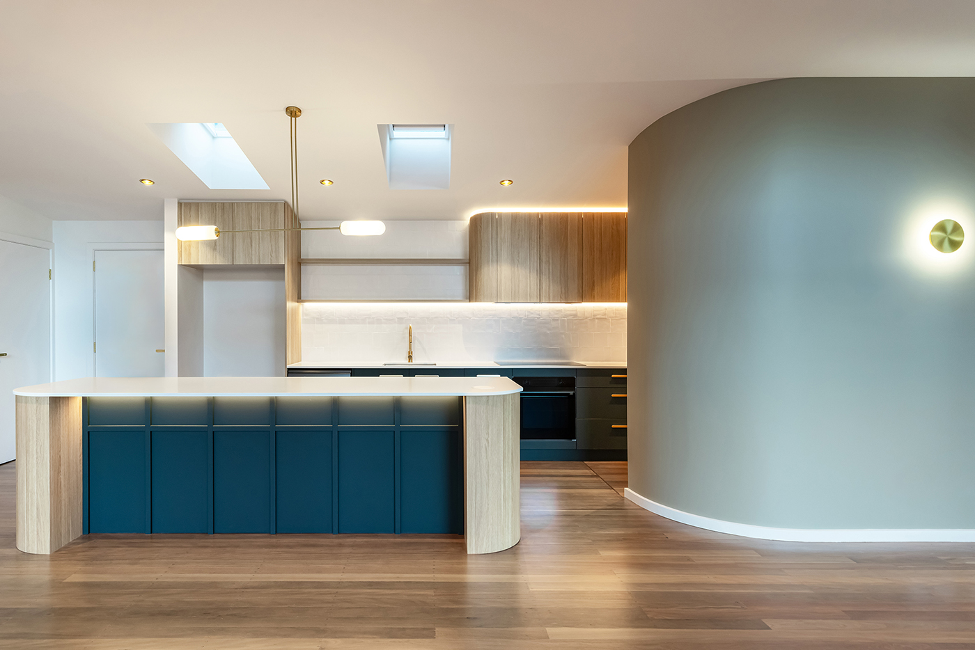 Modern kitchen with blue lower cabinets, light wood upper cabinets, white countertops, two skylights, and curved wall with sconce light.