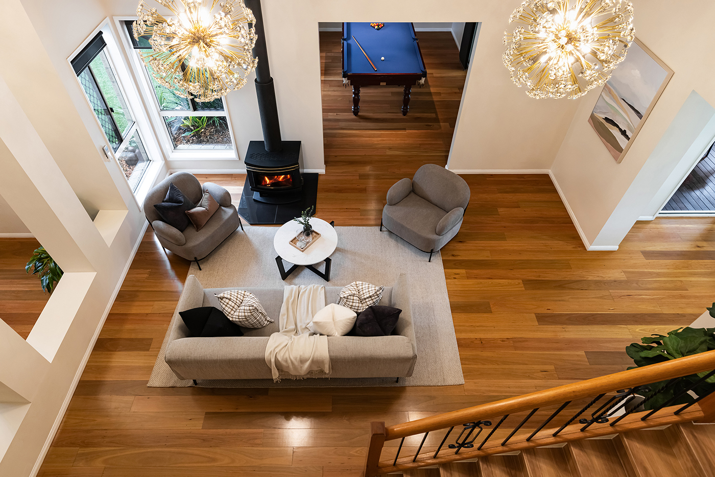 Modern living room with light grey sofa and armchairs around a white round coffee table, wood stove, large windows, wooden floor, chandelier lights, and a billiards table in an adjacent room.