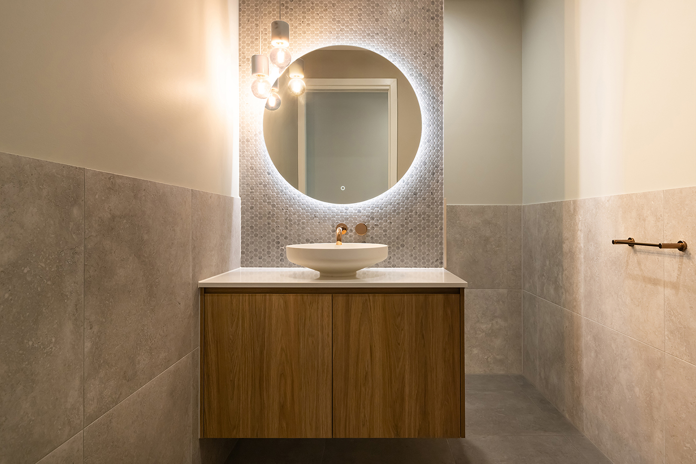 Modern bathroom with a round illuminated mirror, white countertop basin, wooden vanity, and hanging light bulbs.