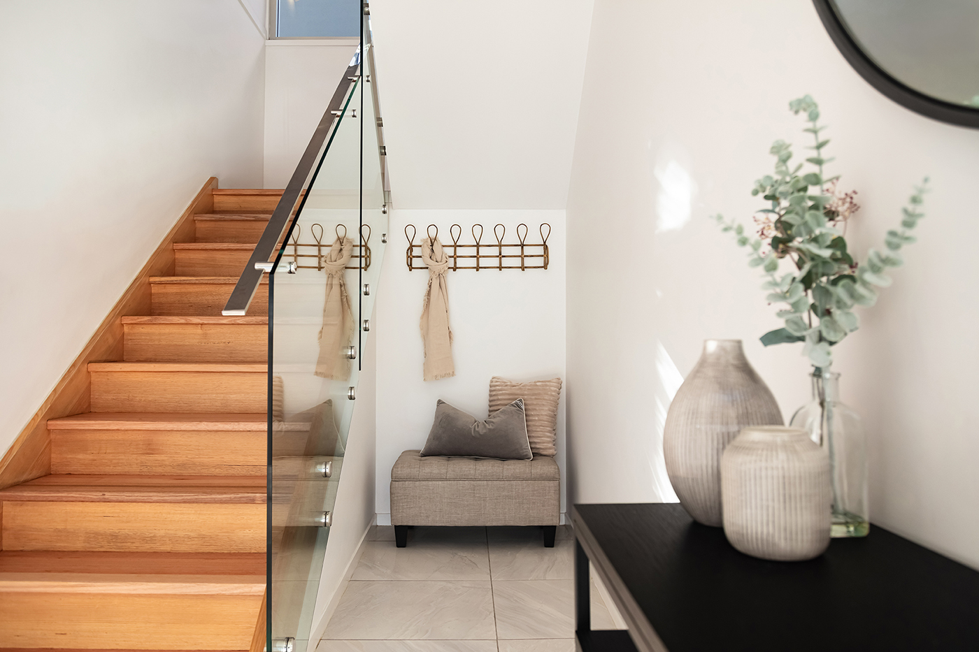 Modern stairway with wooden steps, glass balustrade, beige upholstered bench with pillows, and decorative vases with greenery on a black console table.