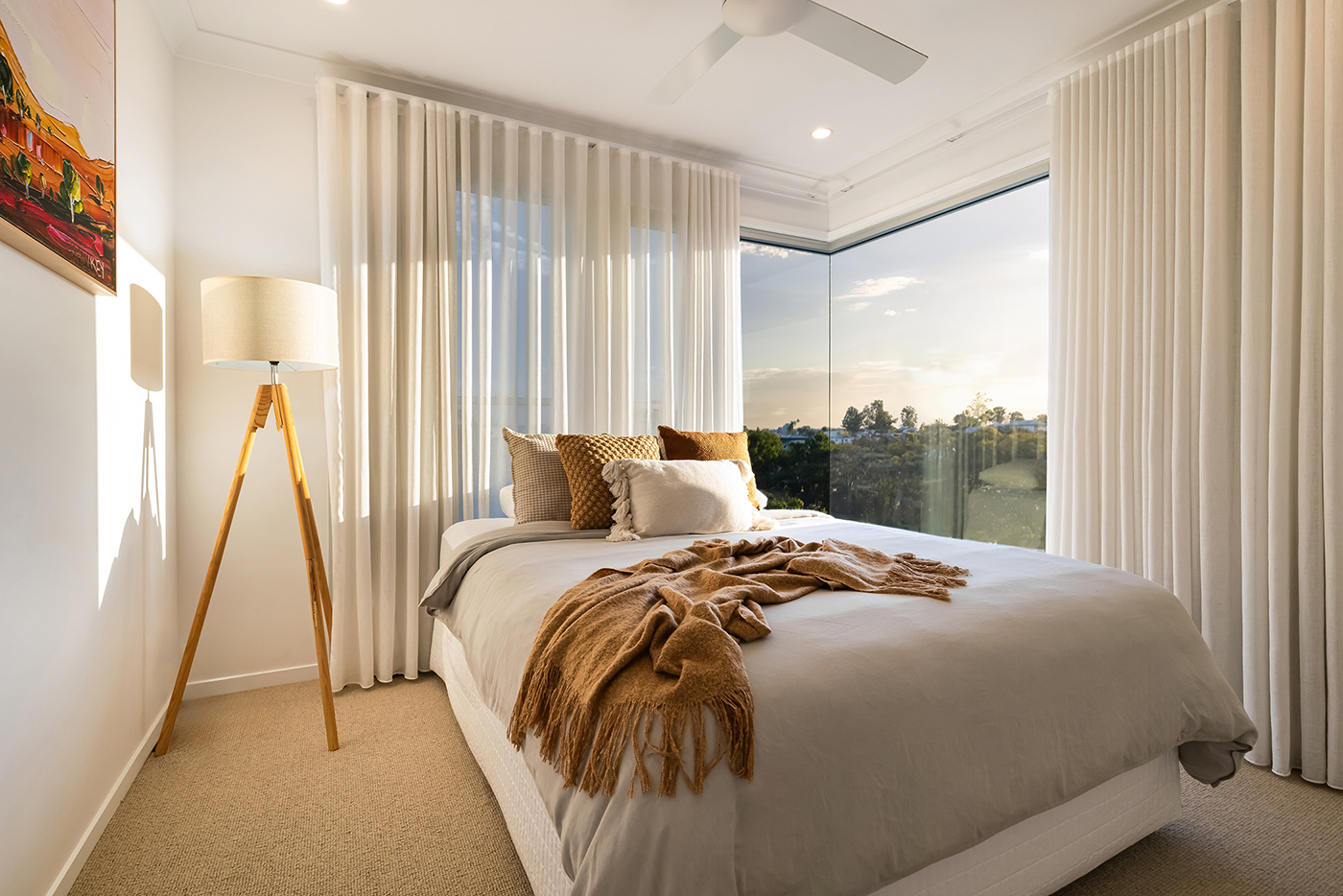 Bright bedroom with a large bed, beige and brown cushions, a brown throw, floor lamp with wooden tripod legs, and a corner window with sheer curtains overlooking trees.