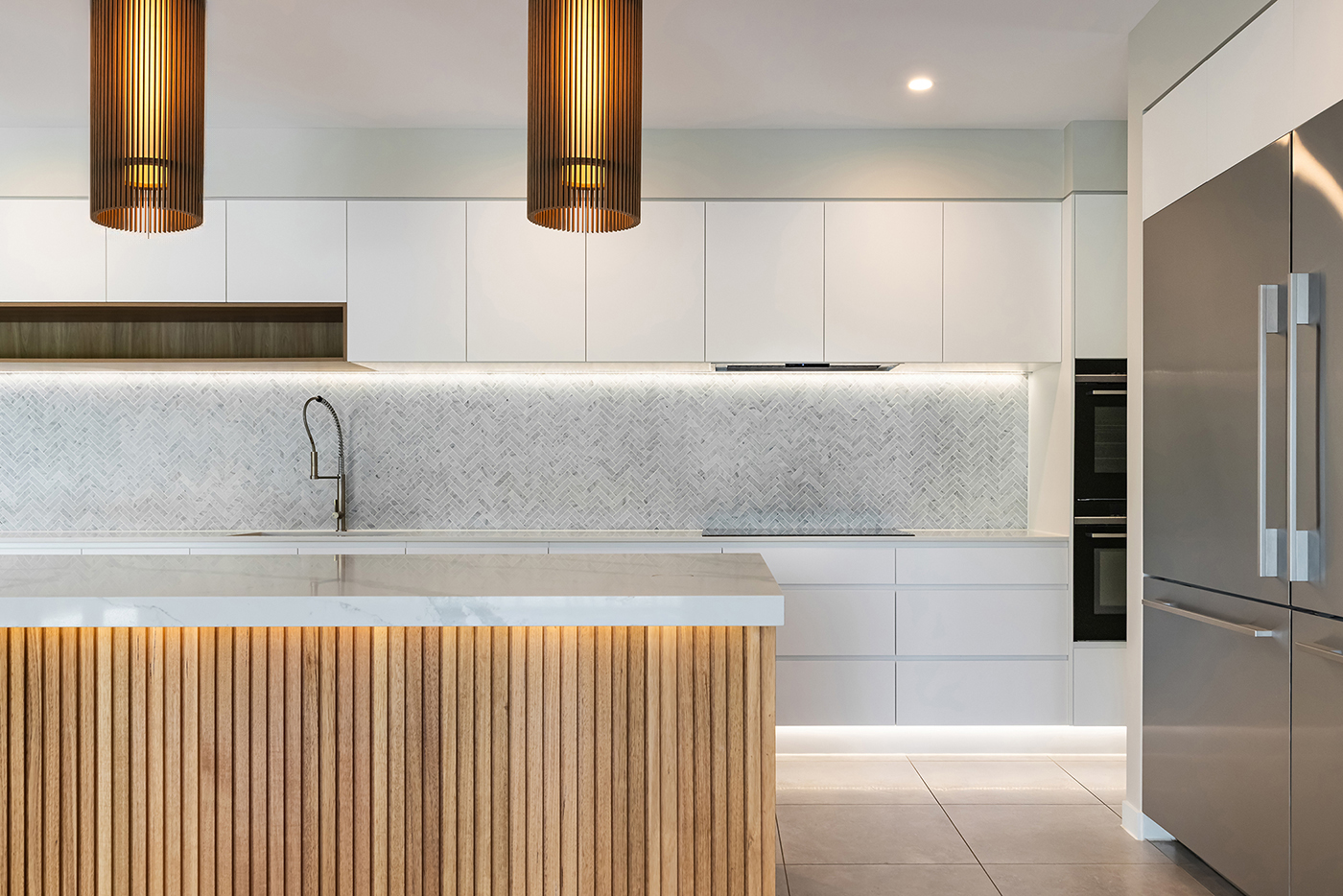 Modern kitchen with white cabinets, marble countertop island with wood paneling, stainless steel refrigerator, and pendant lights.