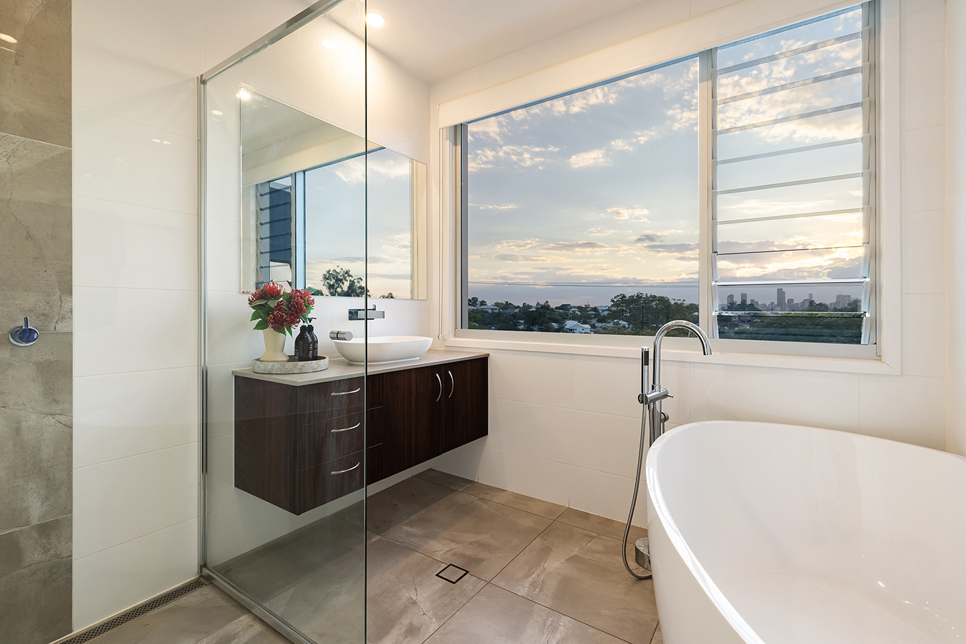 Modern bathroom with freestanding bathtub, glass shower, dark wood vanity with vessel sink, and large window showing a cityscape at sunset.