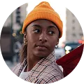 Young woman wearing an orange knit hat and plaid shirt, looking to her left with blurred city lights in the background.