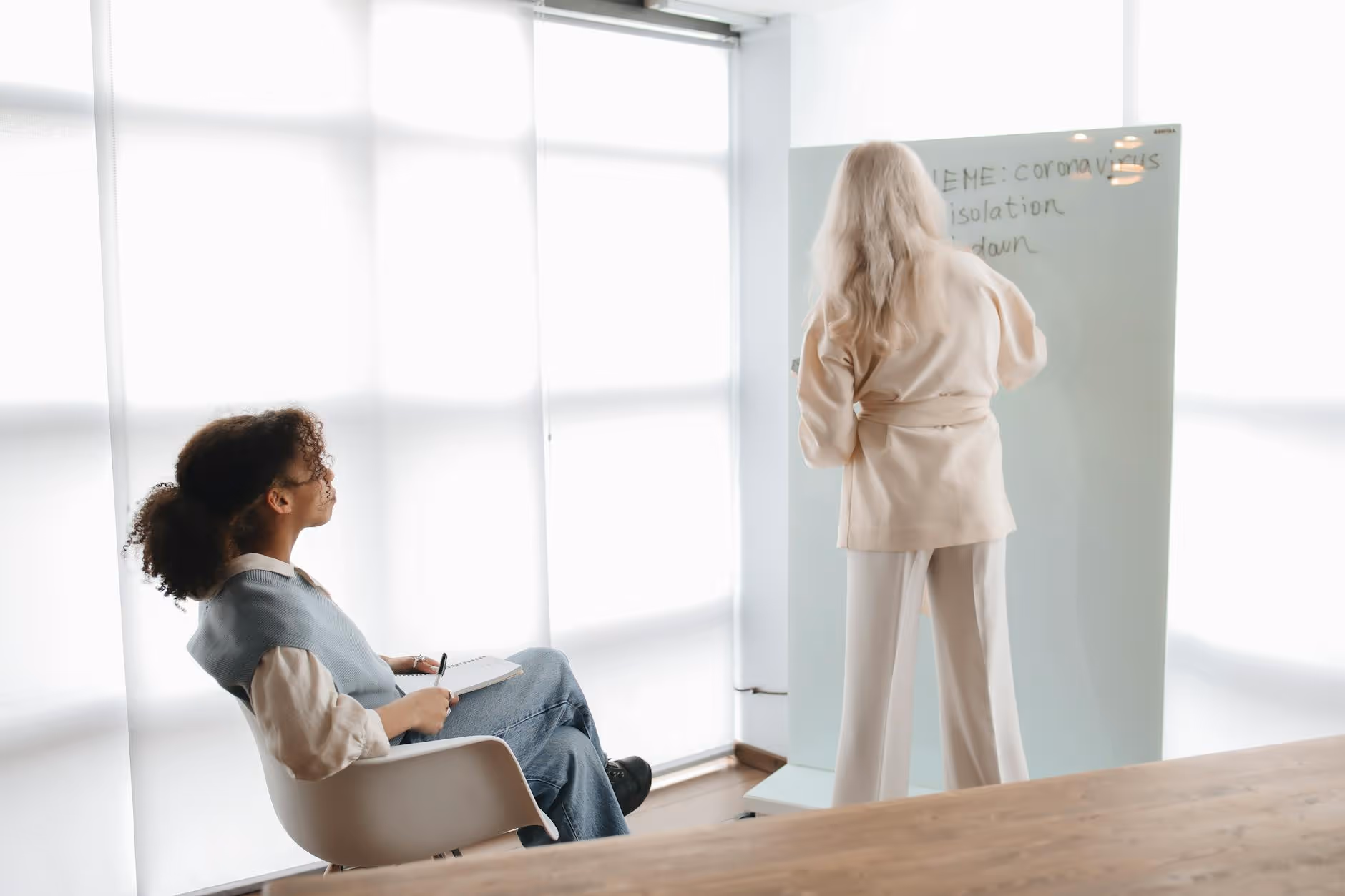 Woman in beige suit writing on whiteboard with words related to coronavirus, while another woman with curly hair sits and takes notes.
