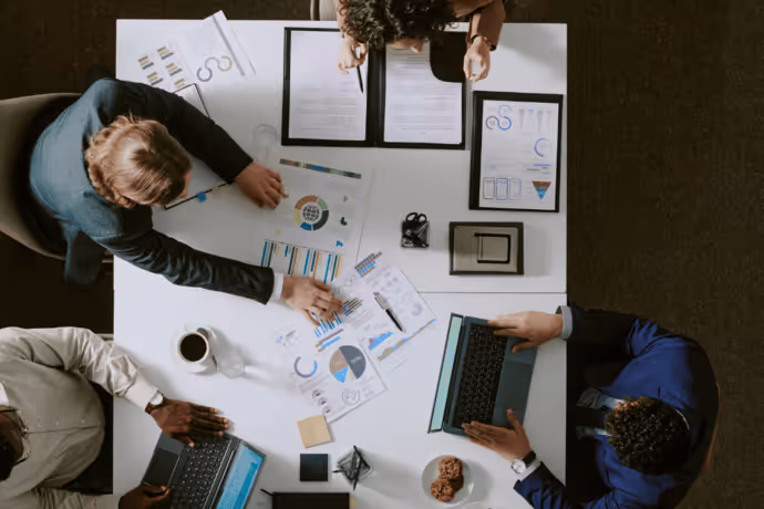 Overhead view of four people working together at a table with laptops, charts, graphs, and a cup of coffee.