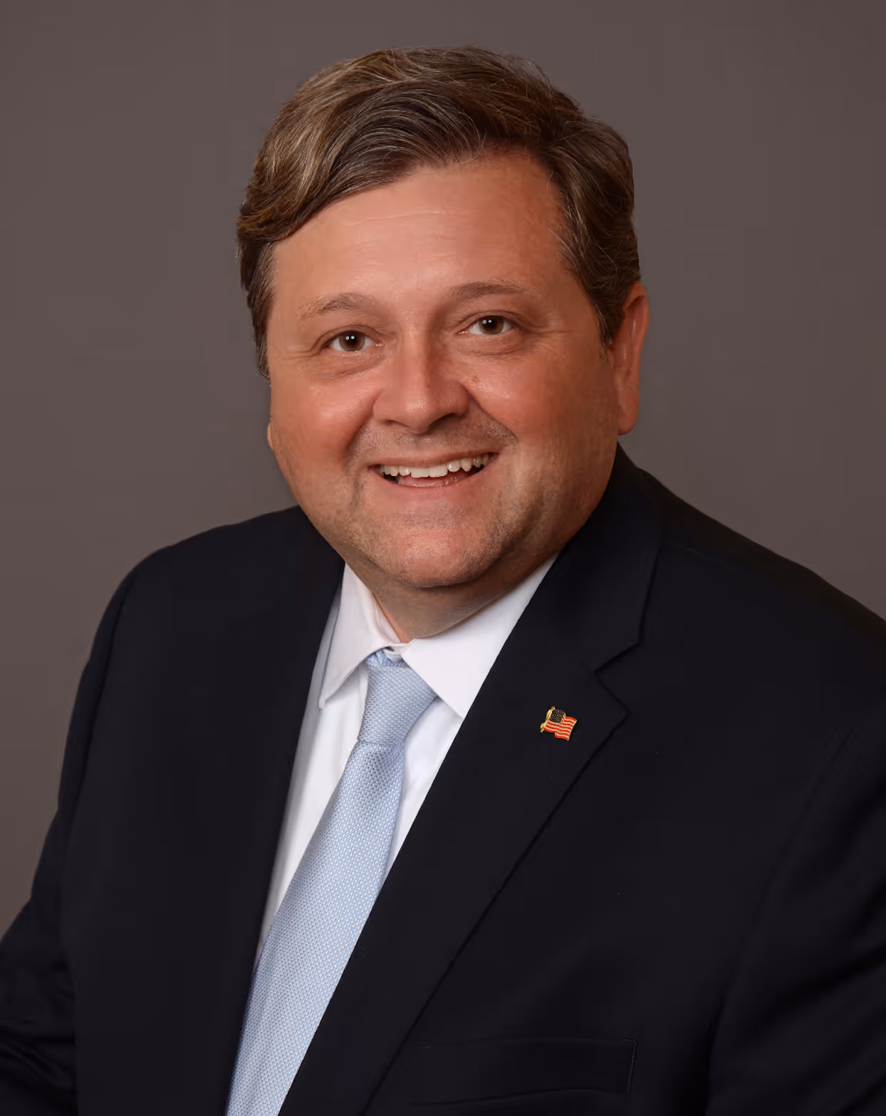 Smiling man with brown hair wearing a dark suit, white shirt, light blue tie, and an American flag pin on the lapel.
