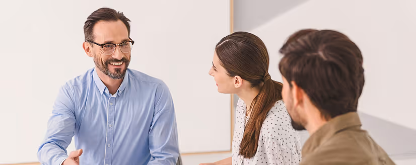 Three people engaged in a discussion, with a smiling man in a blue shirt and glasses speaking to a woman and another man.