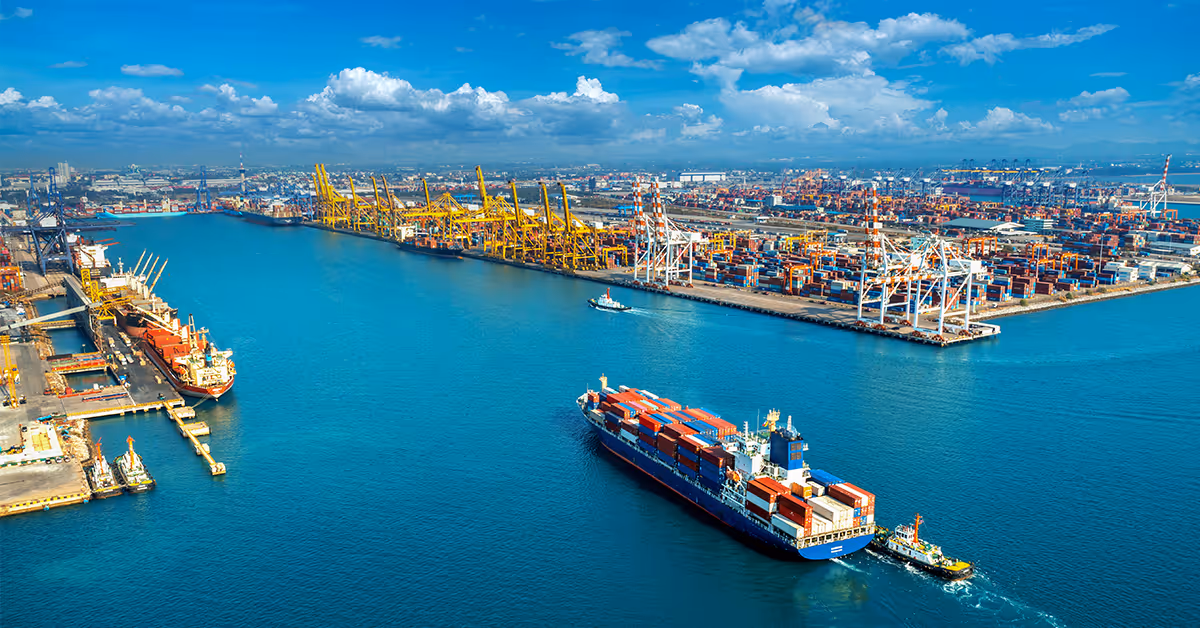 A large cargo ship loaded with colorful containers being guided by a tugboat in a busy port with multiple cranes and container stacks under a blue sky with clouds.