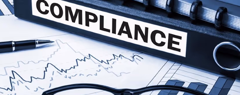 A black binder labeled 'COMPLIANCE' lying on financial charts with a pen and eyeglasses nearby.