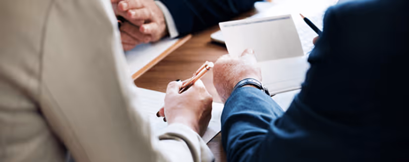 Two people in business attire reviewing documents and taking notes at a wooden table.