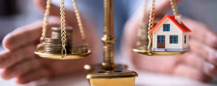 Balance scale comparing stacks of coins and a small house model with hands in the background.