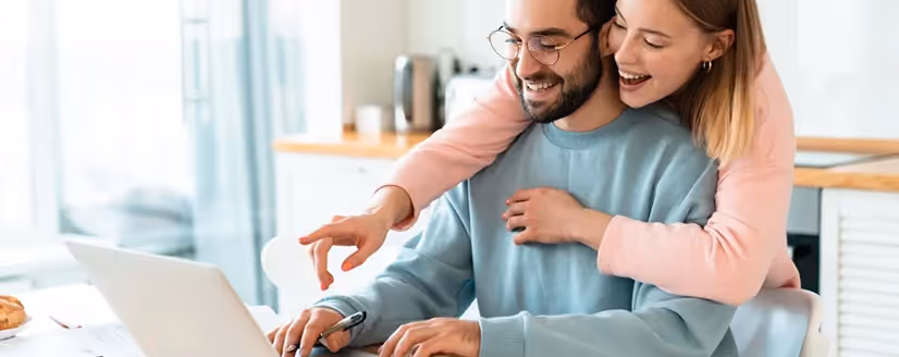 Smiling couple working together on a laptop at home with the woman hugging the man from behind.