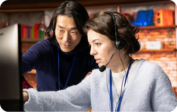 A man and a woman wearing headsets collaborate while looking at a computer screen in an office setting.