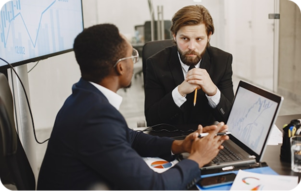 Two businessmen in a meeting discussing financial charts displayed on a laptop and monitor.