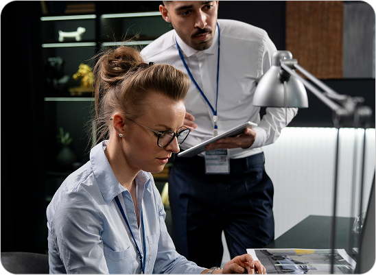 Two office workers, a woman in glasses typing on a keyboard and a man in a white shirt holding a tablet, collaborating in a modern office setting.
