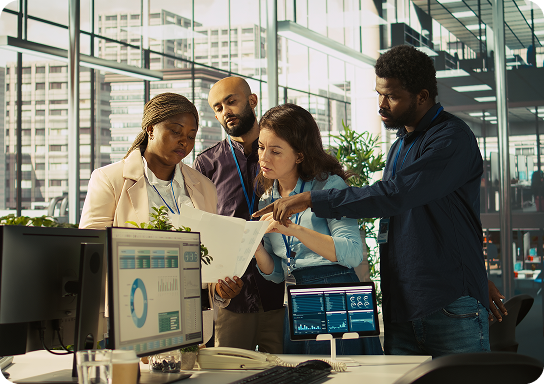 Four diverse coworkers in an office reviewing documents together near computer monitors displaying charts and graphs.