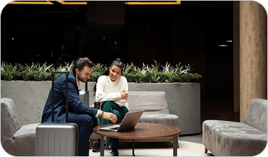 Two business professionals seated on gray sofas looking at a laptop on a round wooden table in a modern lounge area.