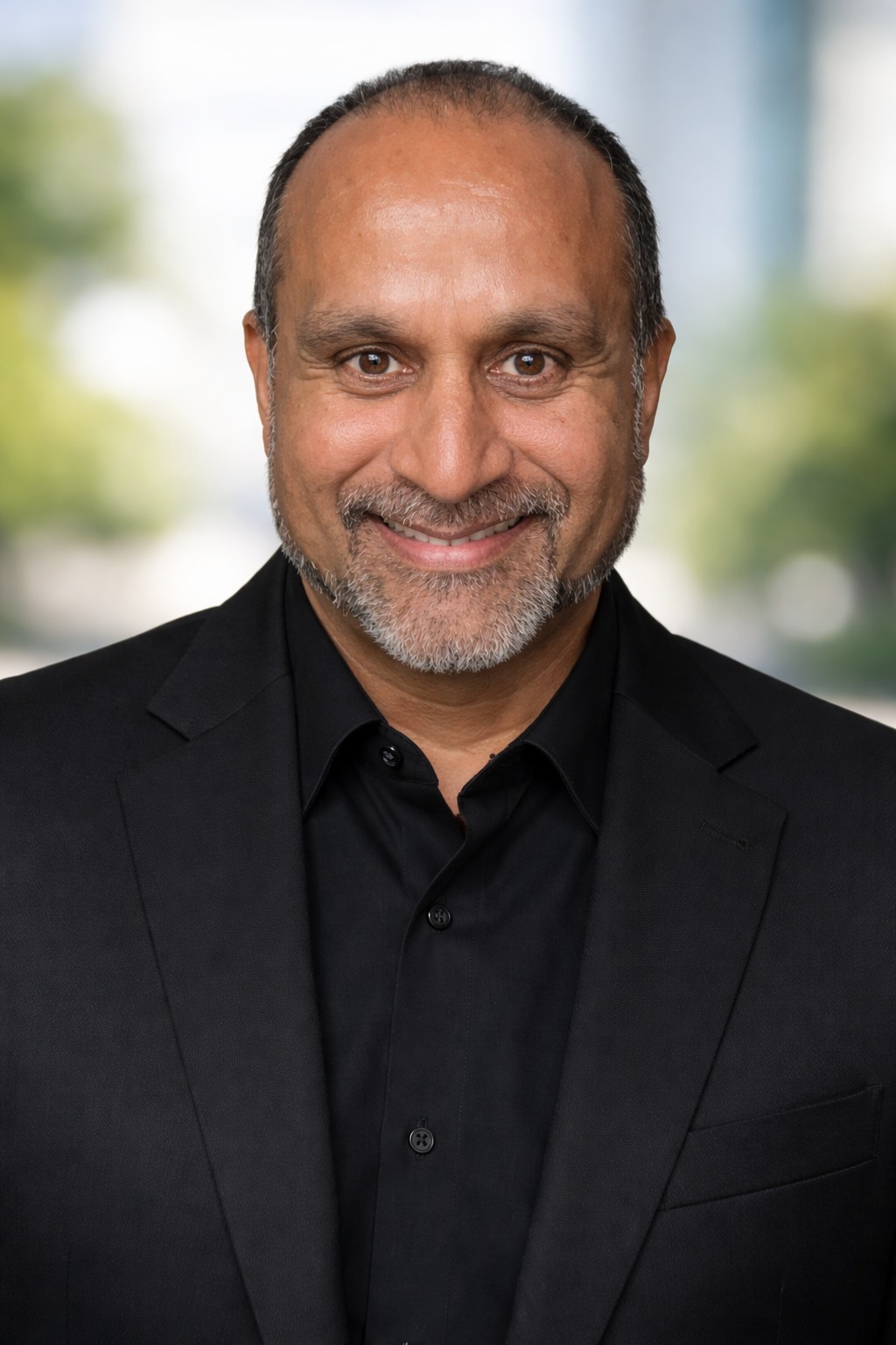 Smiling middle-aged man with a trimmed beard wearing a black suit and shirt against a soft-focused outdoor background.