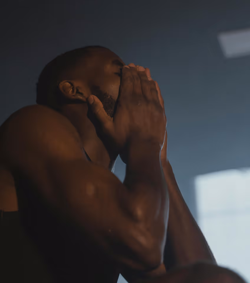 Muscular man in a black tank top with hands clasped near his face in a dimly lit room.