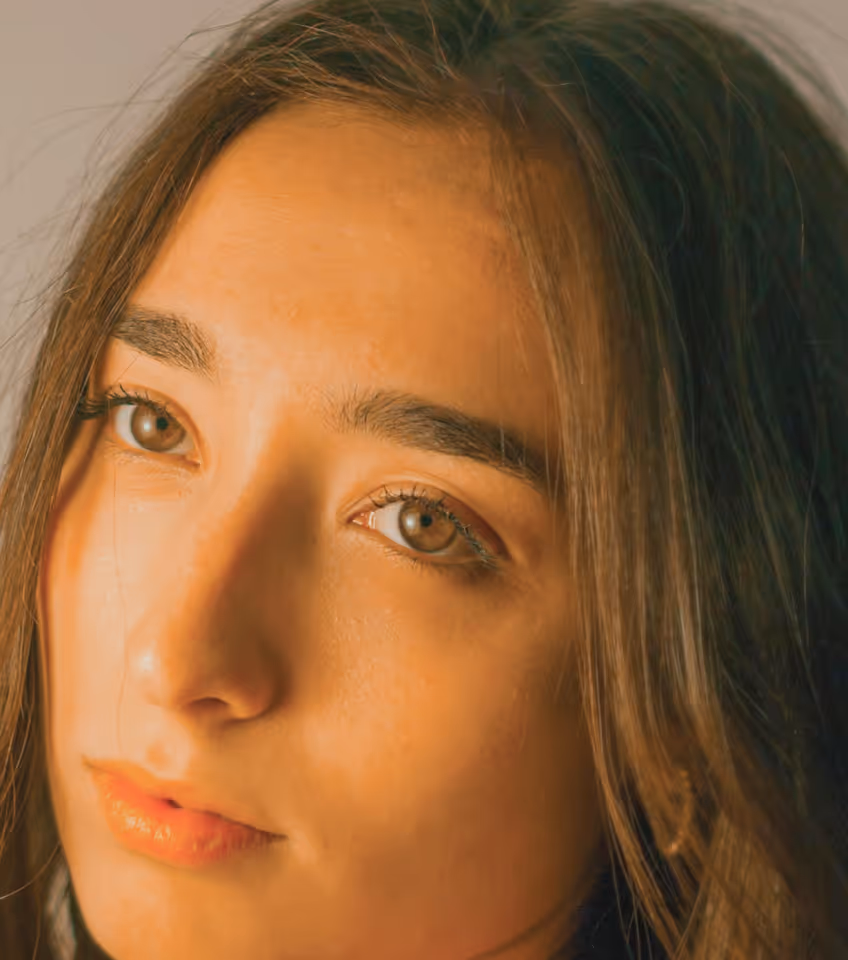 Close-up of a young woman with light brown eyes and long dark hair, softly lit with a warm tone.