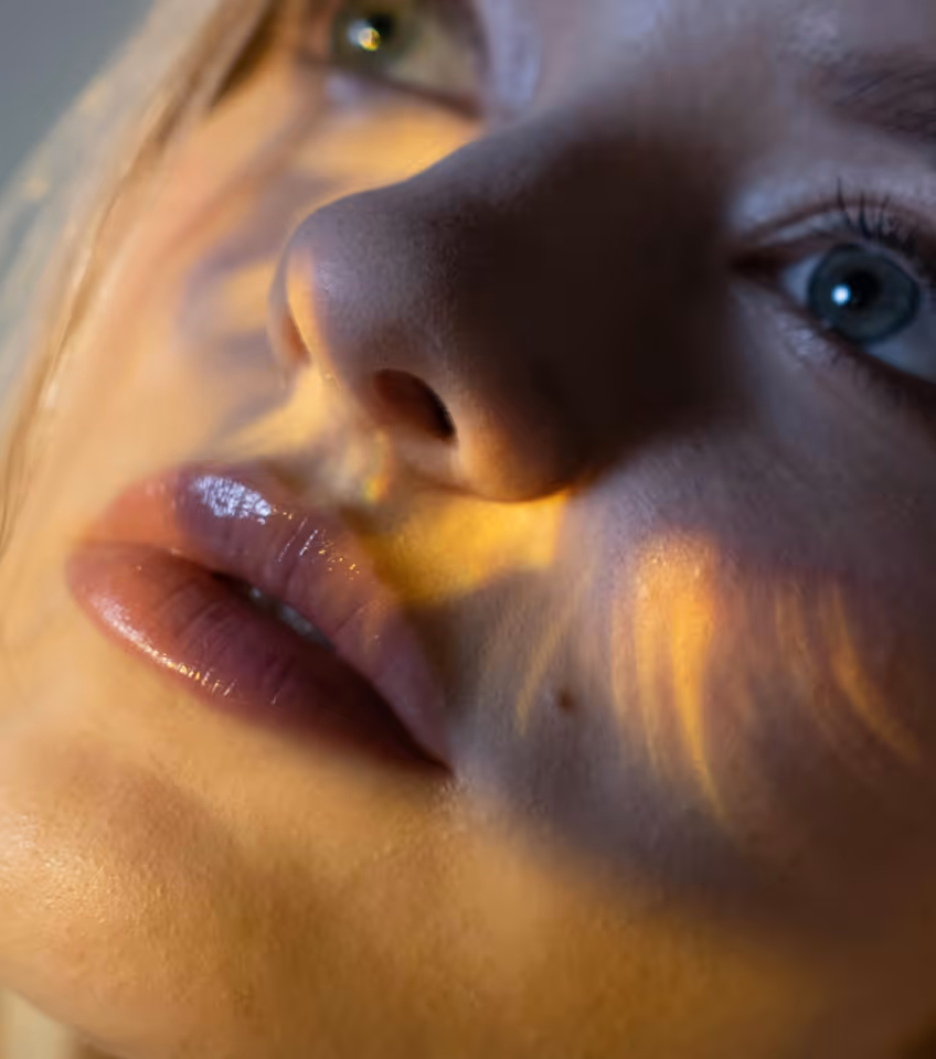 Close-up of a person's face with blue eyes and glossy lips illuminated by warm, soft light patterns.