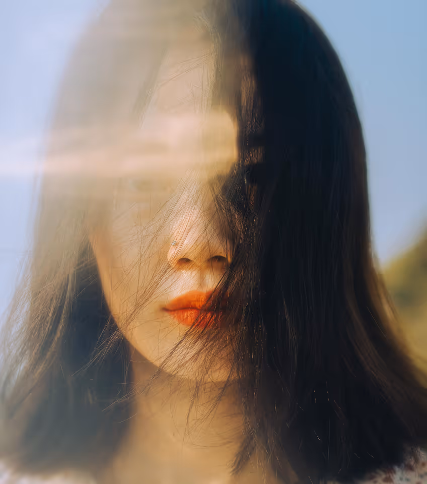 Close-up of a woman with long dark hair partially covering her face and red lipstick, with sunlight streaks across the image.
