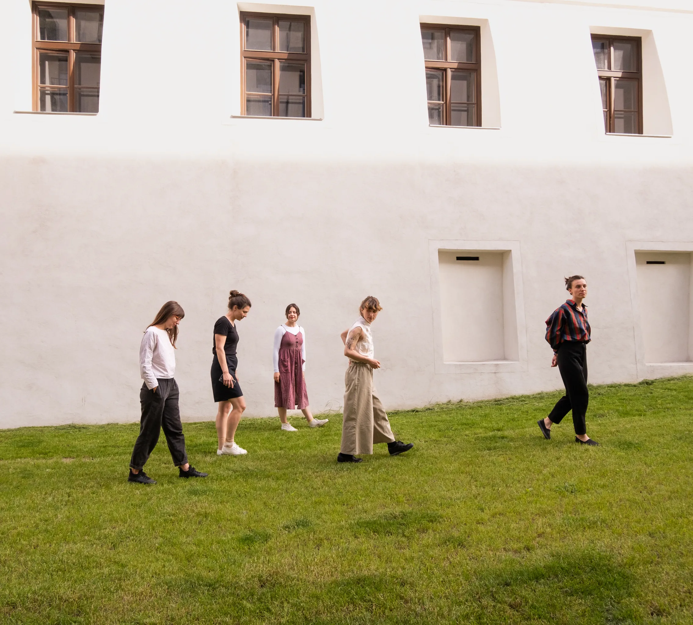 Päť mladých žien kráča po zelenej tráve pred historickou budovou s hnedými drevenými oknami a bielou fasádou.