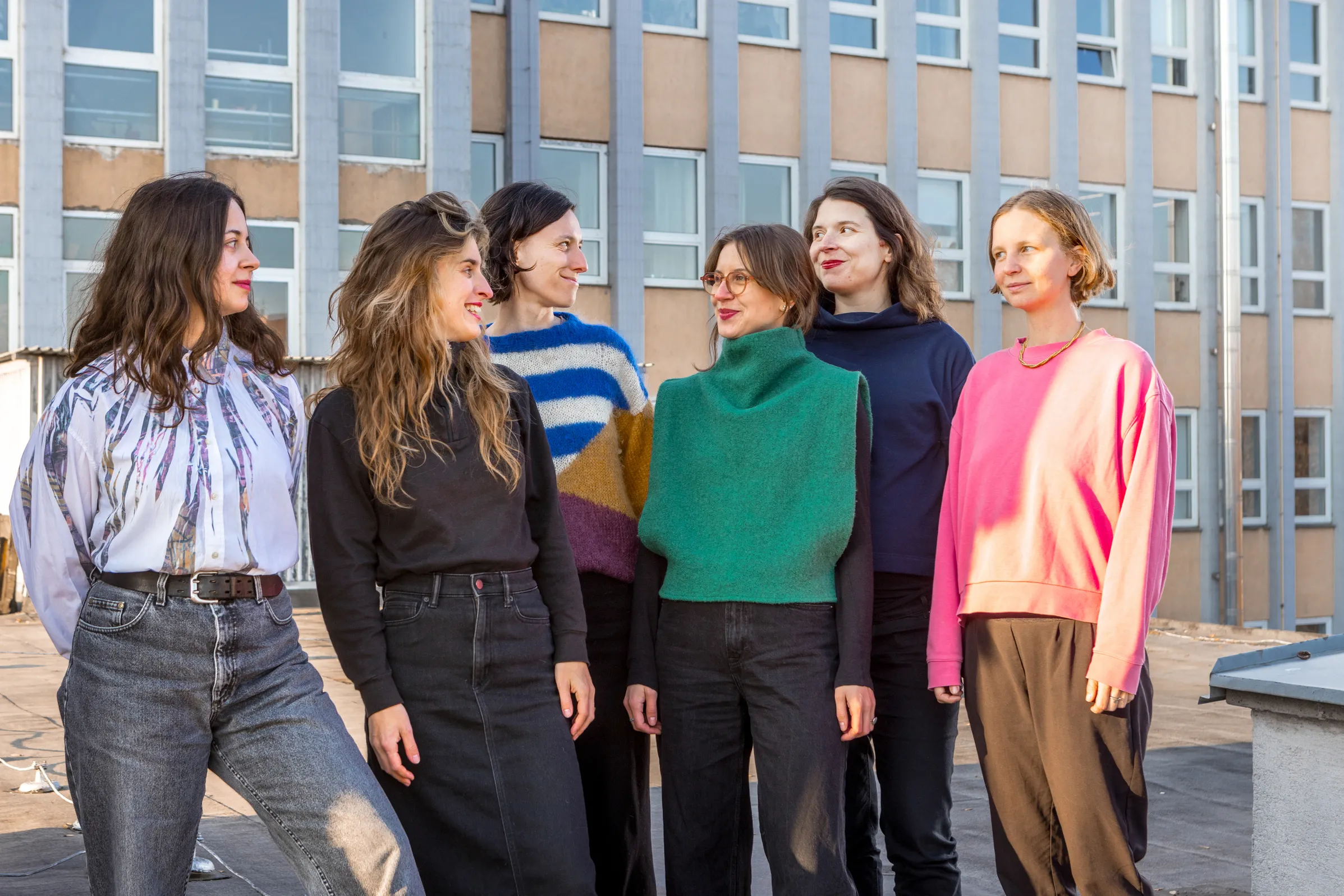 Šesť žien stojí vonku pred budovou, usmievajú sa a rozprávajú sa medzi sebou.