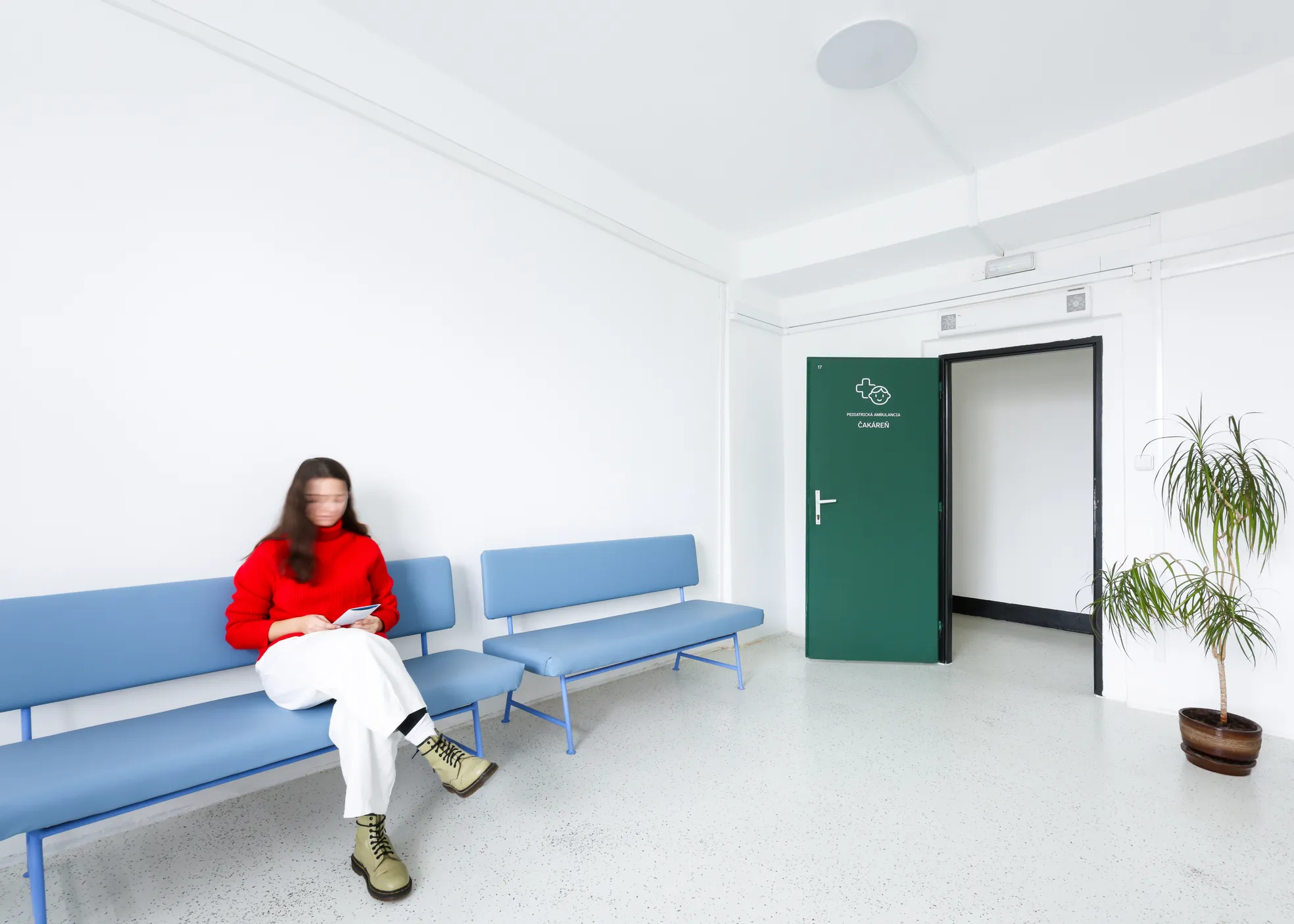 Žena v červenom svetri sedí na modrej lavičke v čakárni s otvorenými zelenými dverami označenými 'Pediatrická ambulancia'.