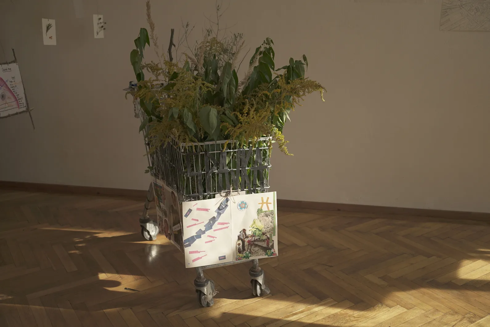 Nákupný vozík so zelenými listami a žltými kvetmi na drevenej podlahe v miestnosti so svetlými stenami.