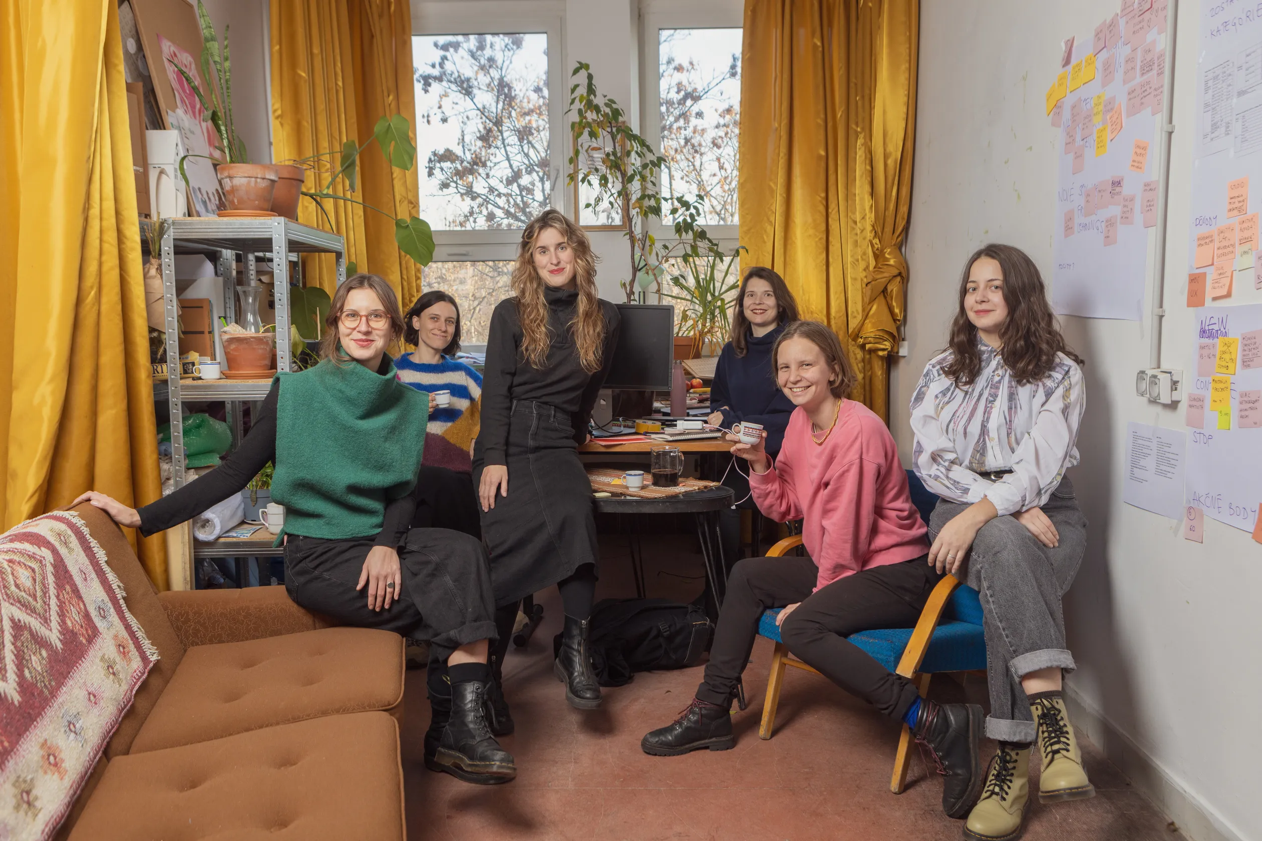 Šesť žien sedí a usmieva sa v útulnej kancelárskej miestnosti s oknami, žltými závesmi, rastlinami a tabuľou so samolepkami na stene.