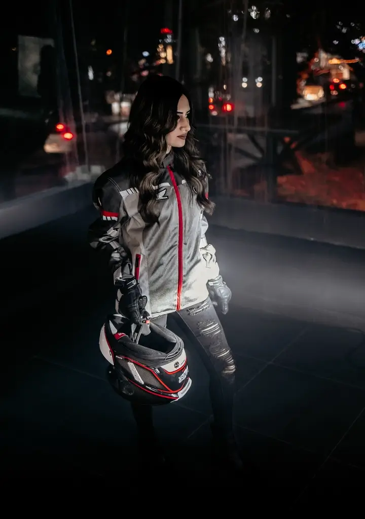 Woman wearing a gray and red riding jacket holding a black and red motorcycle helmet in a dimly lit urban setting.