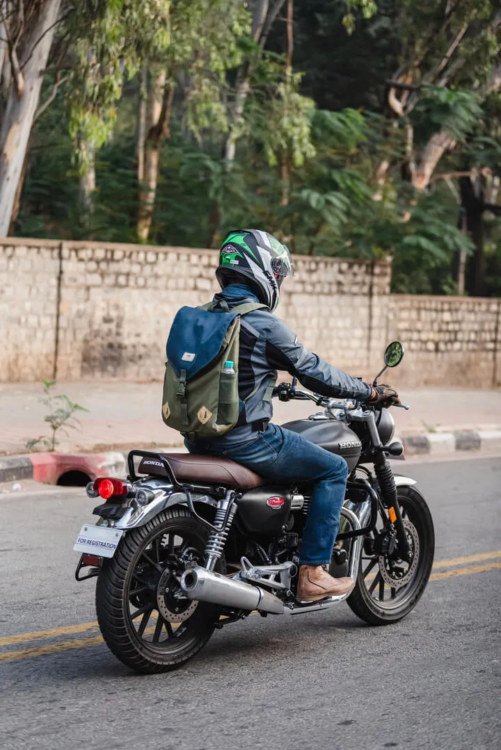 Motorcyclist wearing a helmet and backpack riding a black Honda motorcycle on a street lined with trees and a stone wall.