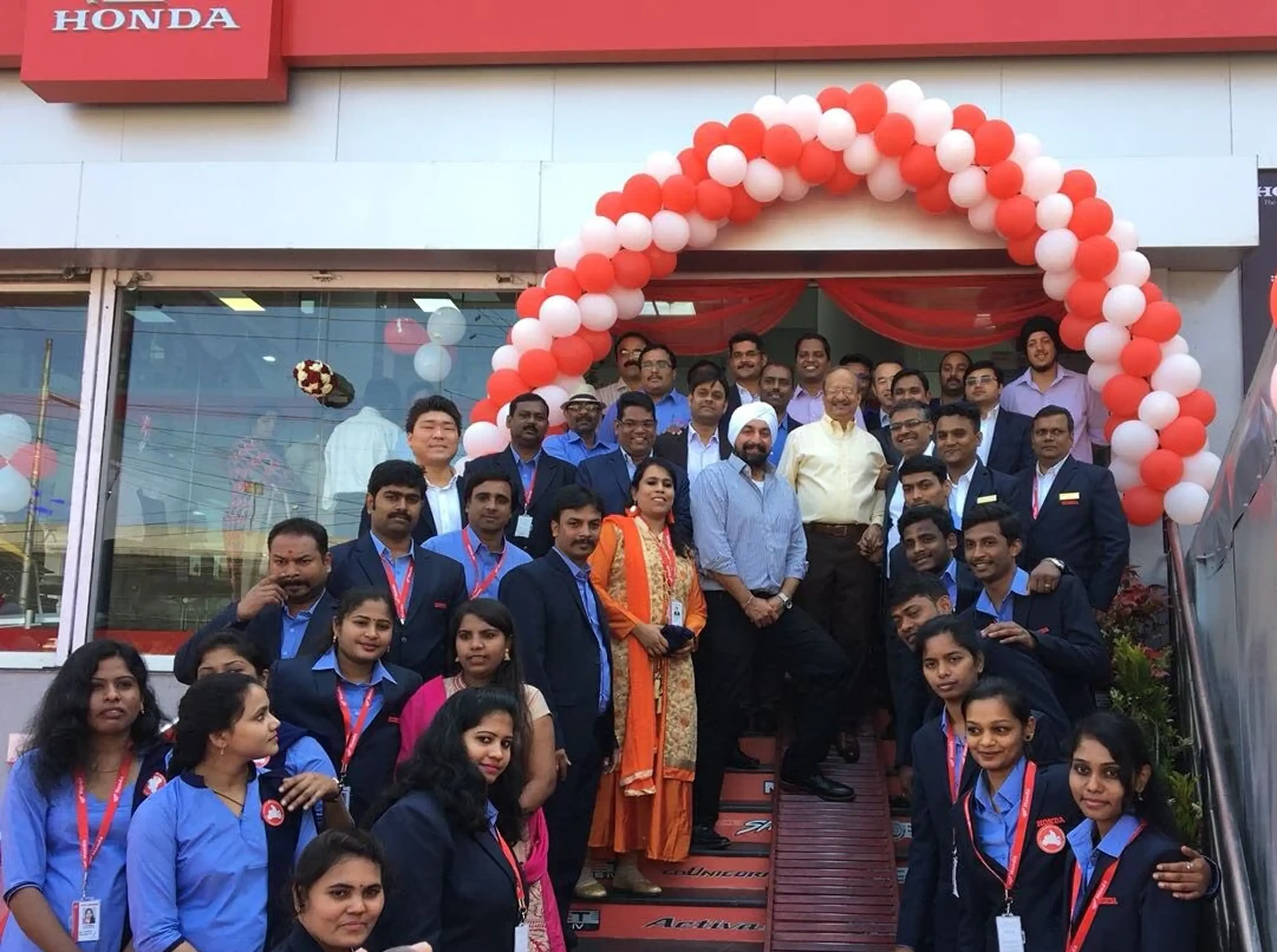 Group of men and women posing under a red and white balloon arch in front of a Honda dealership.