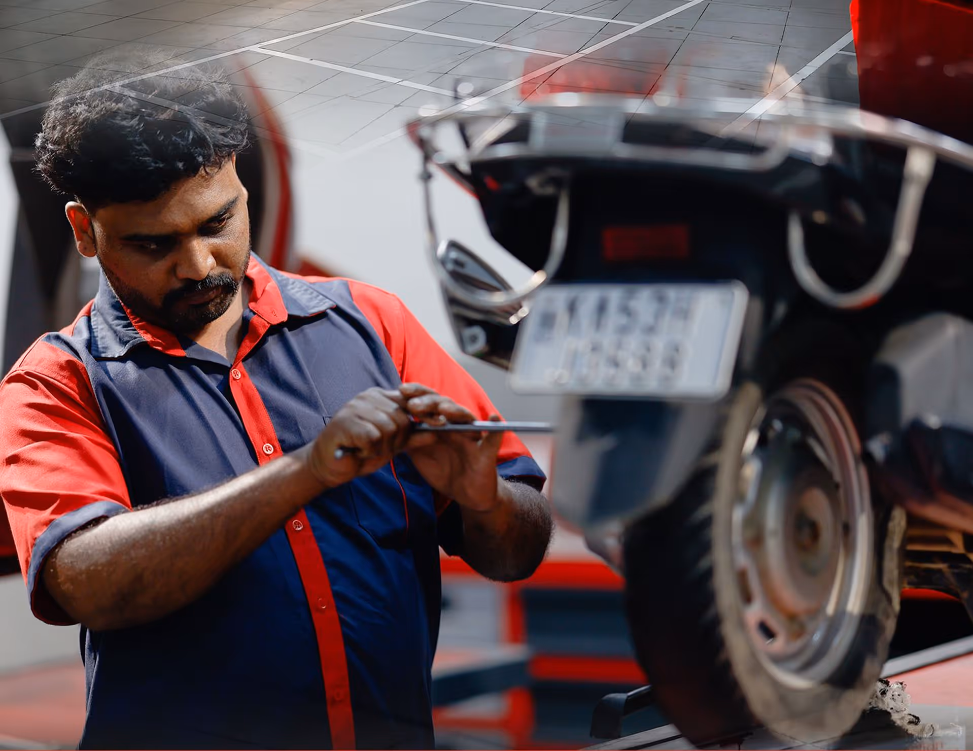 Mechanic in a red and navy uniform repairing a scooter wheel with a screwdriver in a workshop.