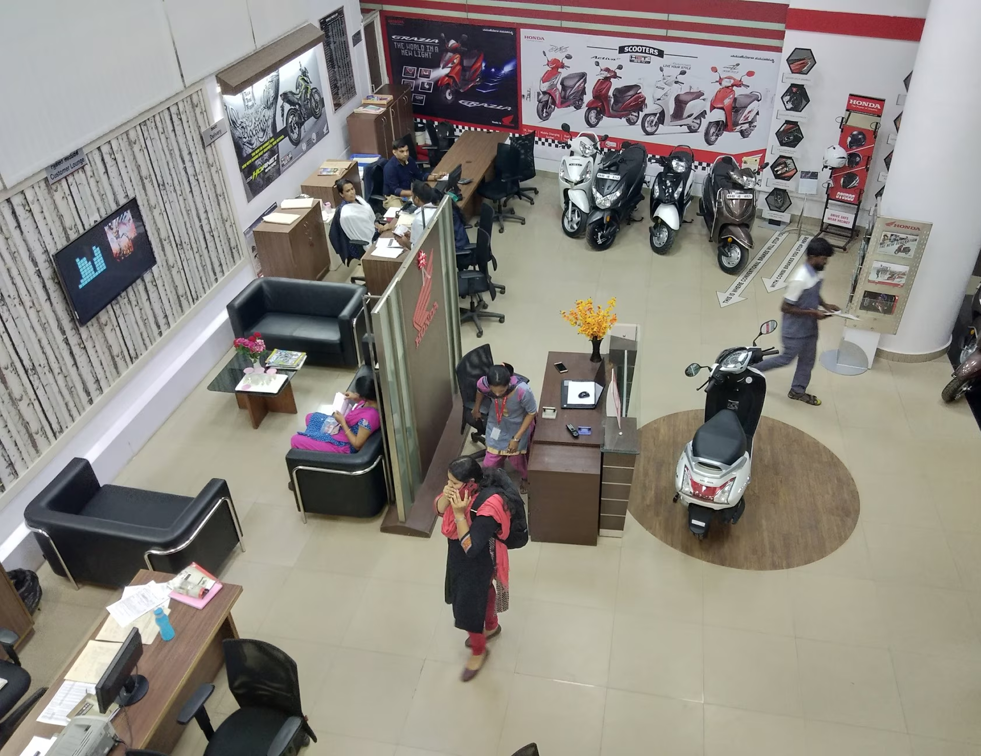 Interior of a Honda showroom with scooters displayed, customers seated, and staff working at desks.