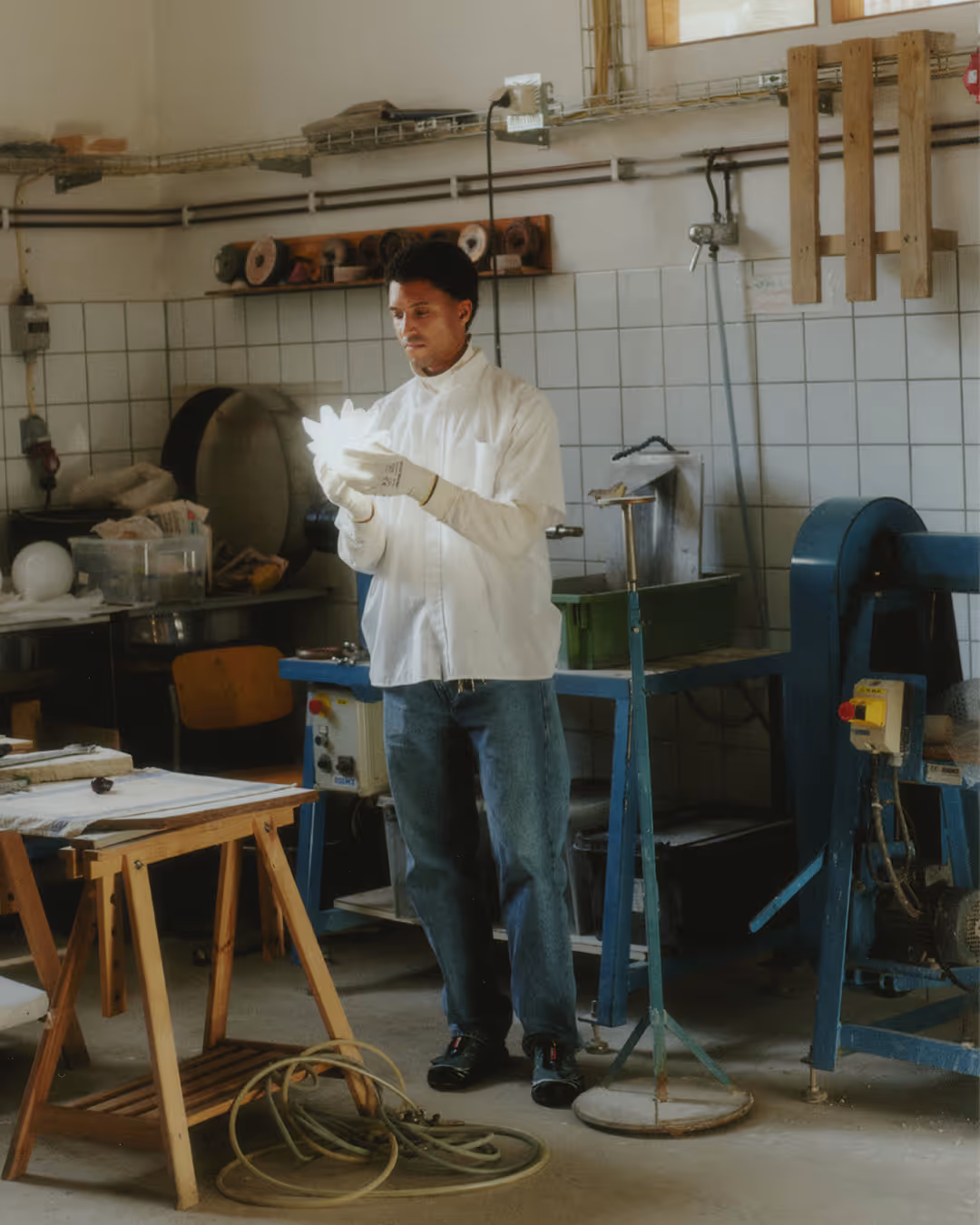 Simon Skinner wearing white gloves and lab coat inspecting a white object in a workshop with machinery and wooden table.