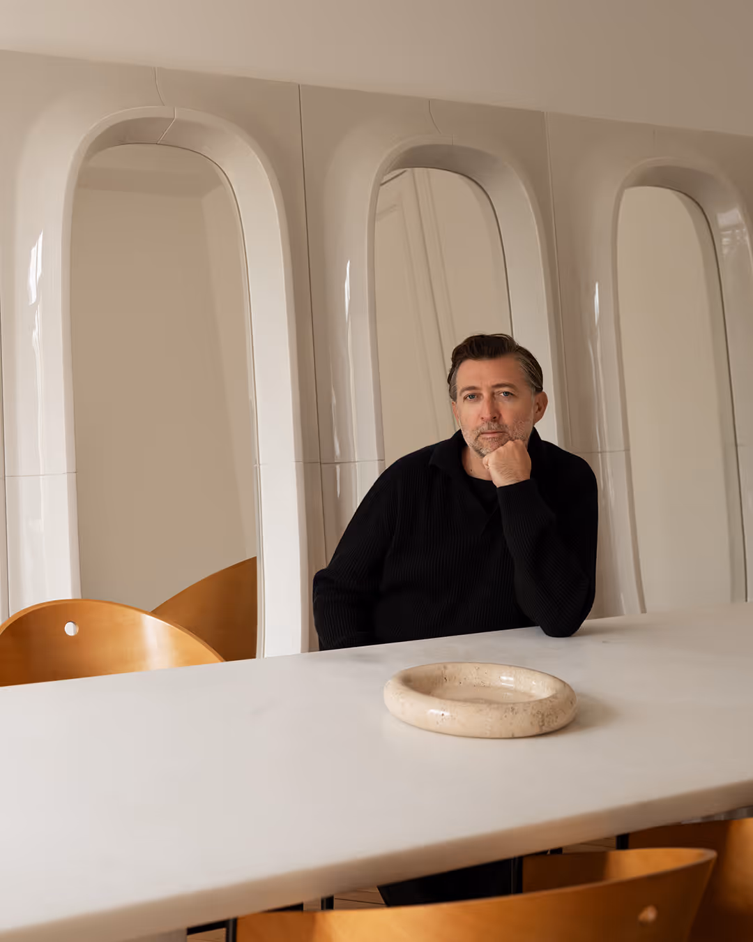 Willo Perron in a black sweater sitting at a white table with modern golden chairs and arched wall panels behind him.