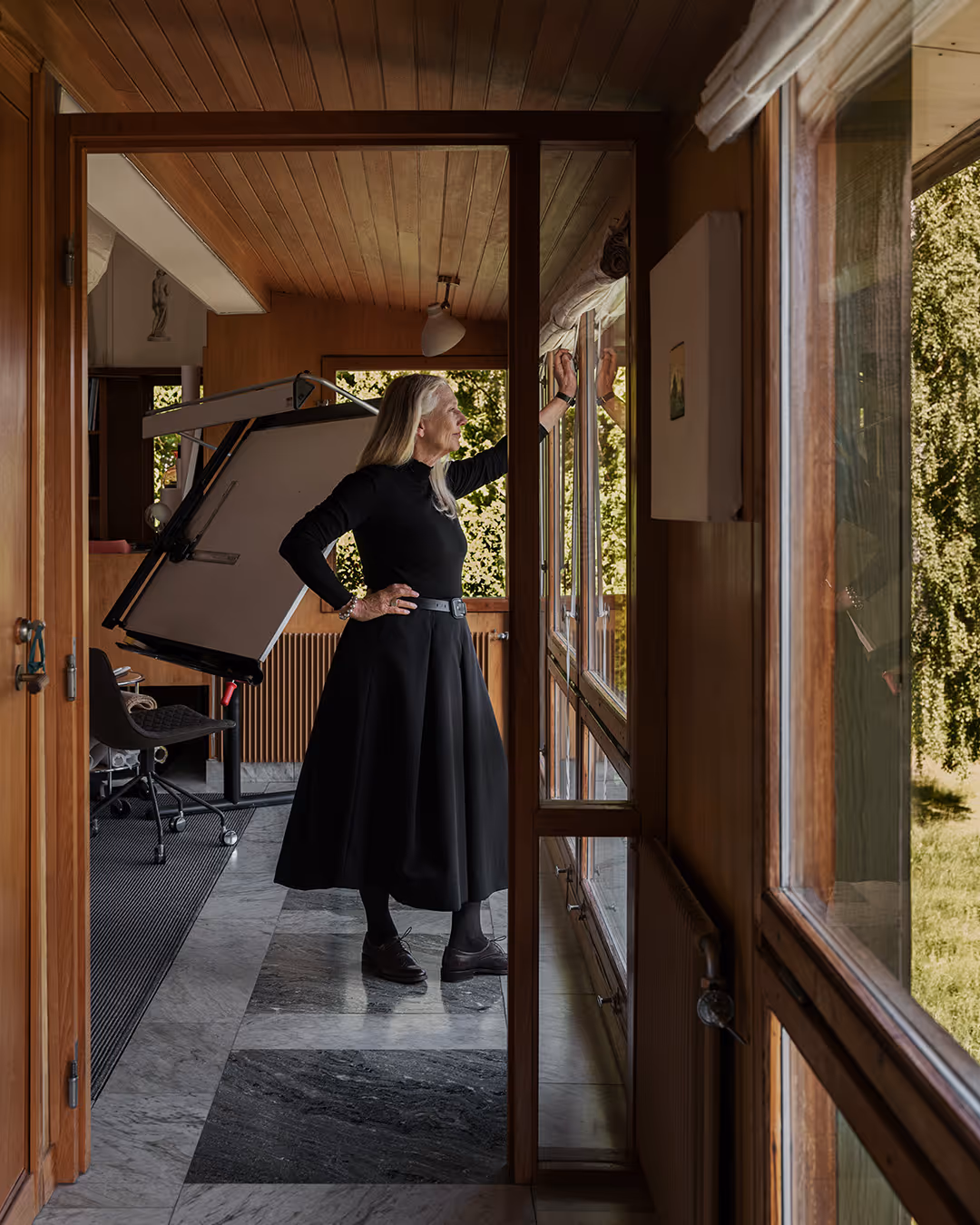 Gunilla Allard dressed in black standing indoors, adjusting window blinds with a drawing table and chair visible in the background.