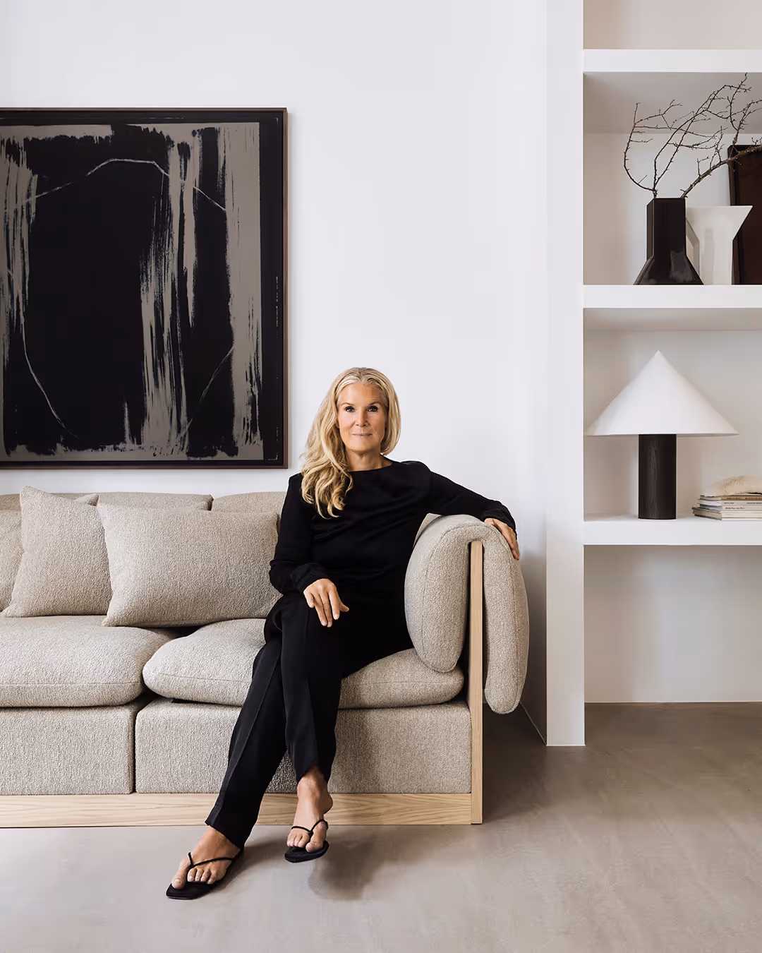 Lotta Agaton in black outfit sitting on a beige modern sofa in a minimalist living room with abstract black and gray painting and white shelves with decor.