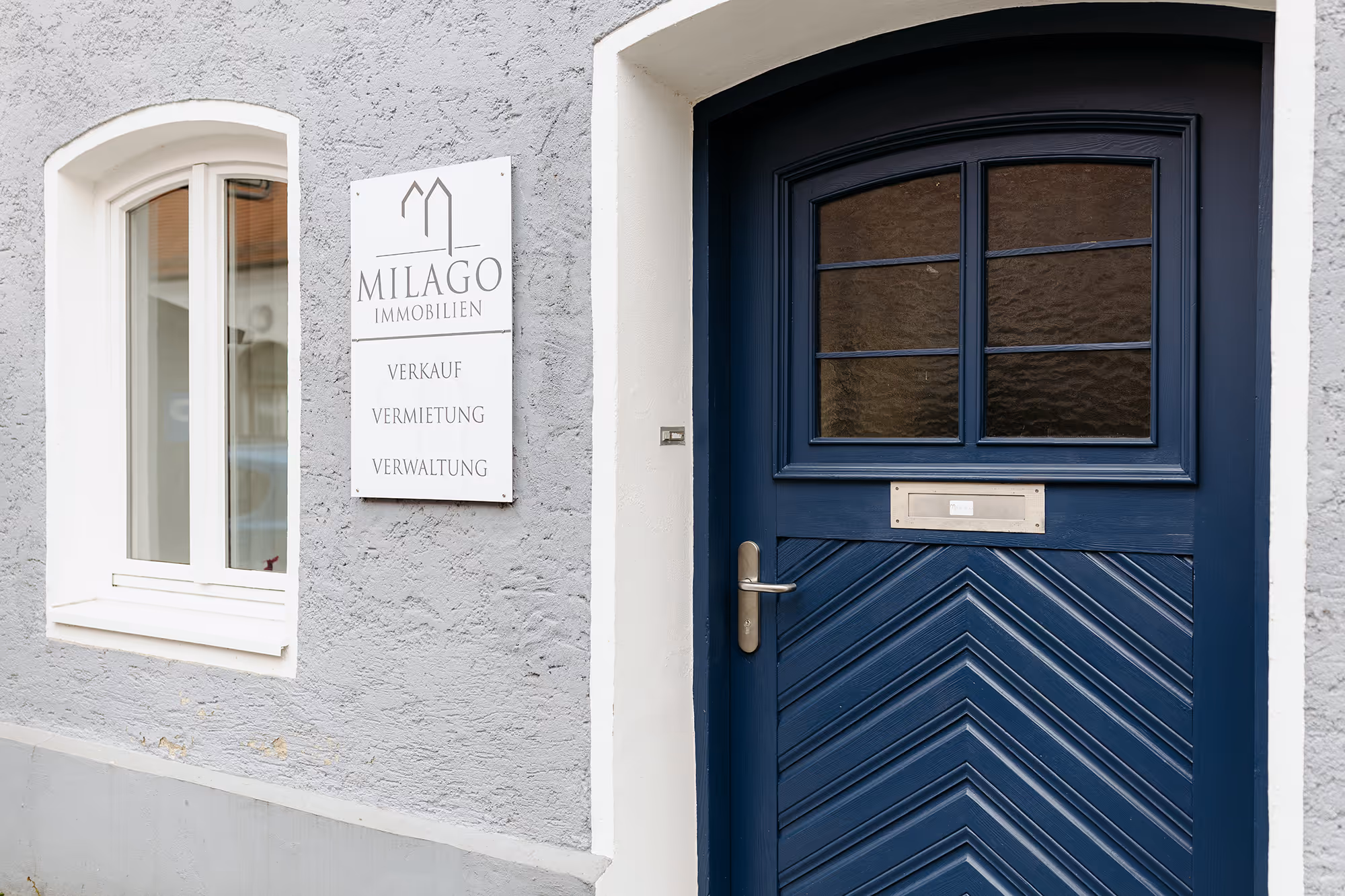 Eingangstür eines Gebäudes mit einem blauen Holztür und einem Schild daneben mit der Aufschrift 'Milago Immobilien Verkauf Vermietung Verwaltung'.