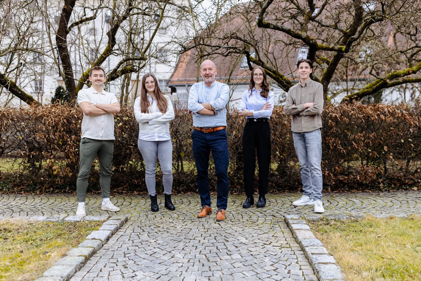Fünf Personen stehen mit verschränkten Armen auf einem gepflasterten Weg in einem Garten im Freien.