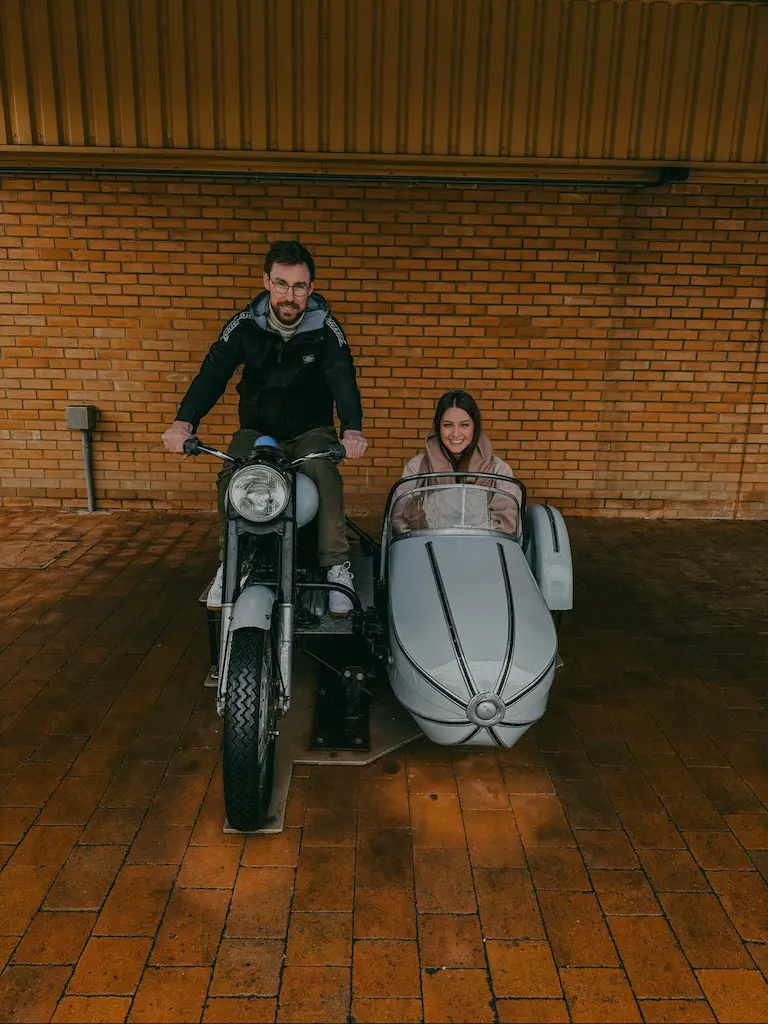Félix sur une moto avec Lisa assise dans un side-car gris souriant, devant un mur en briques orange.
