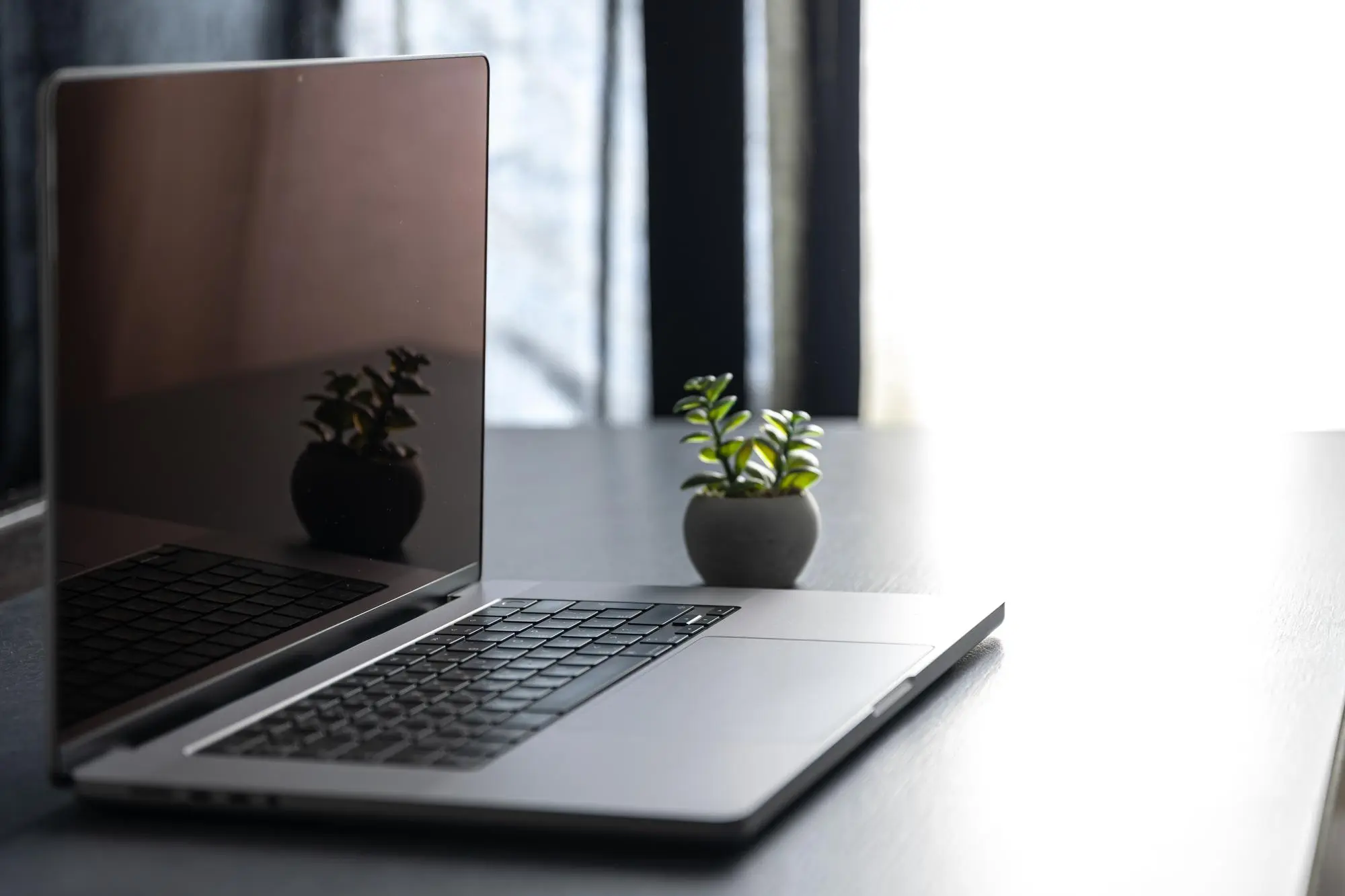 Ordinateur portable ouvert avec un petit pot de plante sur une table devant une fenêtre lumineuse.