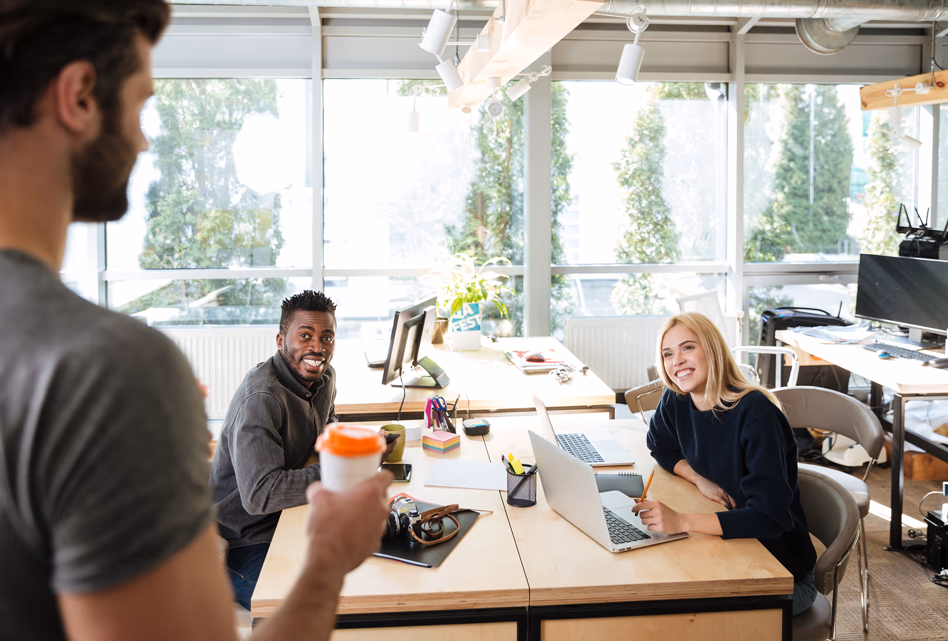 Trois collègues discutent dans un bureau lumineux avec des fenêtres, deux sont assis à une table avec des ordinateurs portables, un debout tenant une tasse.