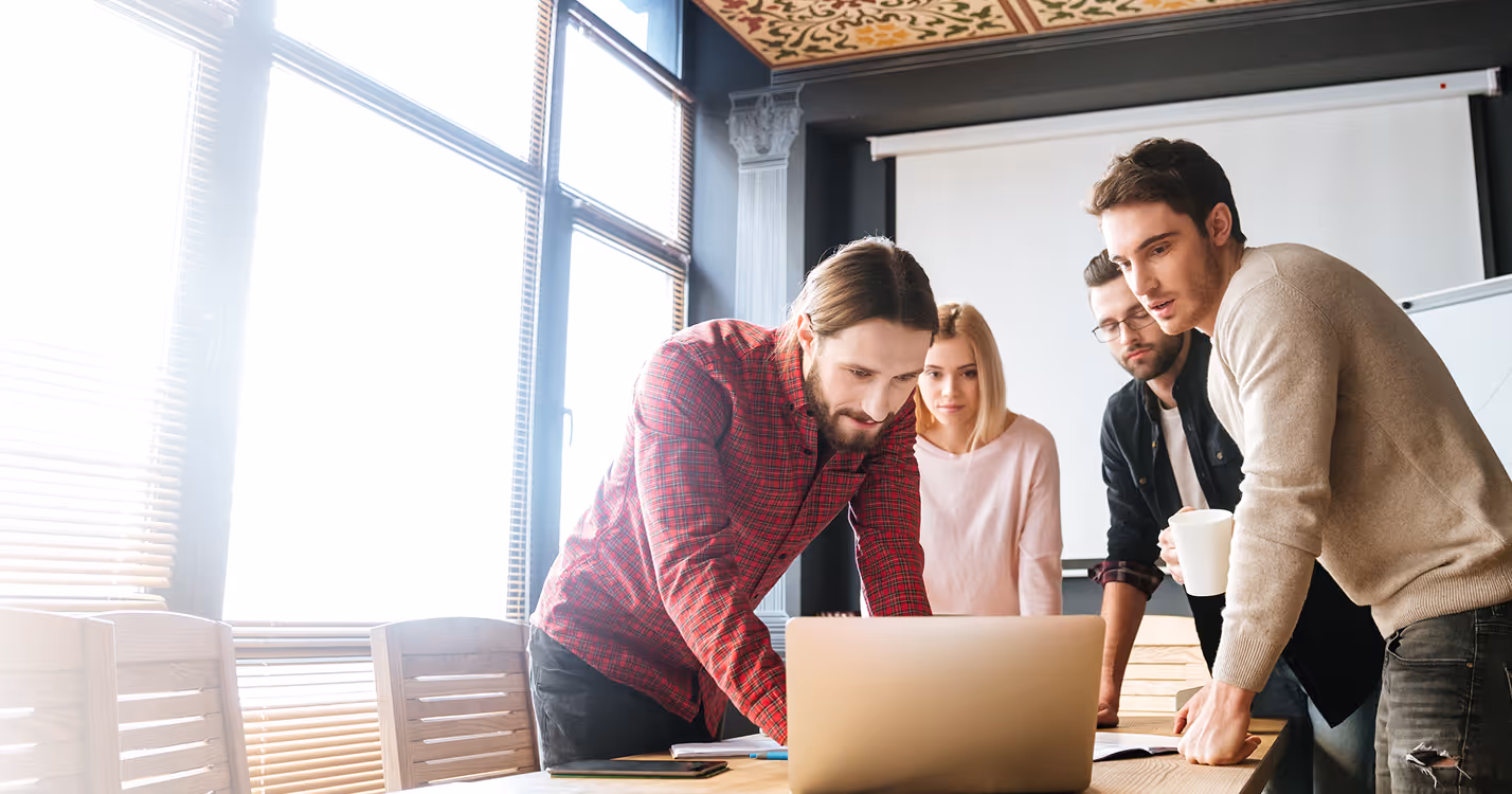 Quatre jeunes professionnels concentrés discutent autour d'un ordinateur portable dans une salle de réunion lumineuse.