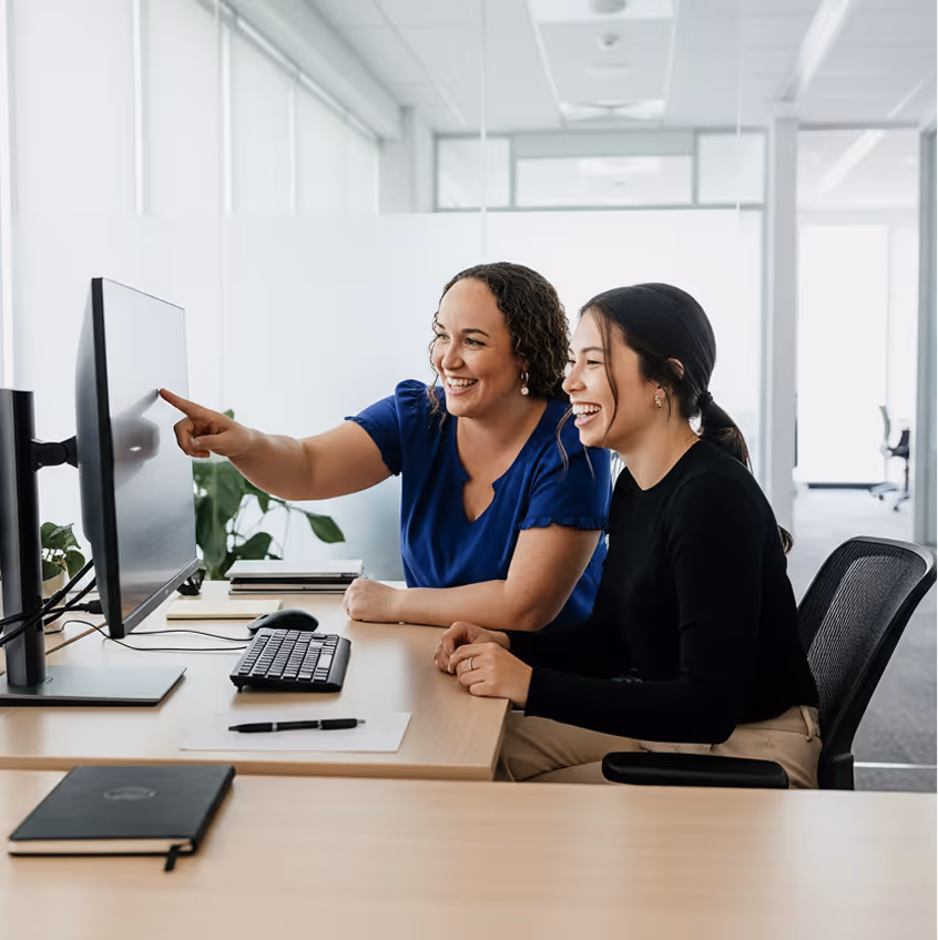Deux femmes souriantes regardent un écran d'ordinateur dans un bureau lumineux, l'une d'elles pointant l'écran.